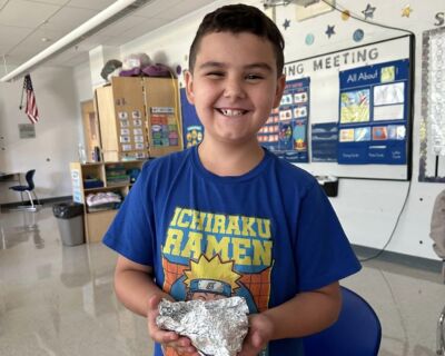 A smiling young boy in class with a blue naruto shirt on holding some aluminum foil